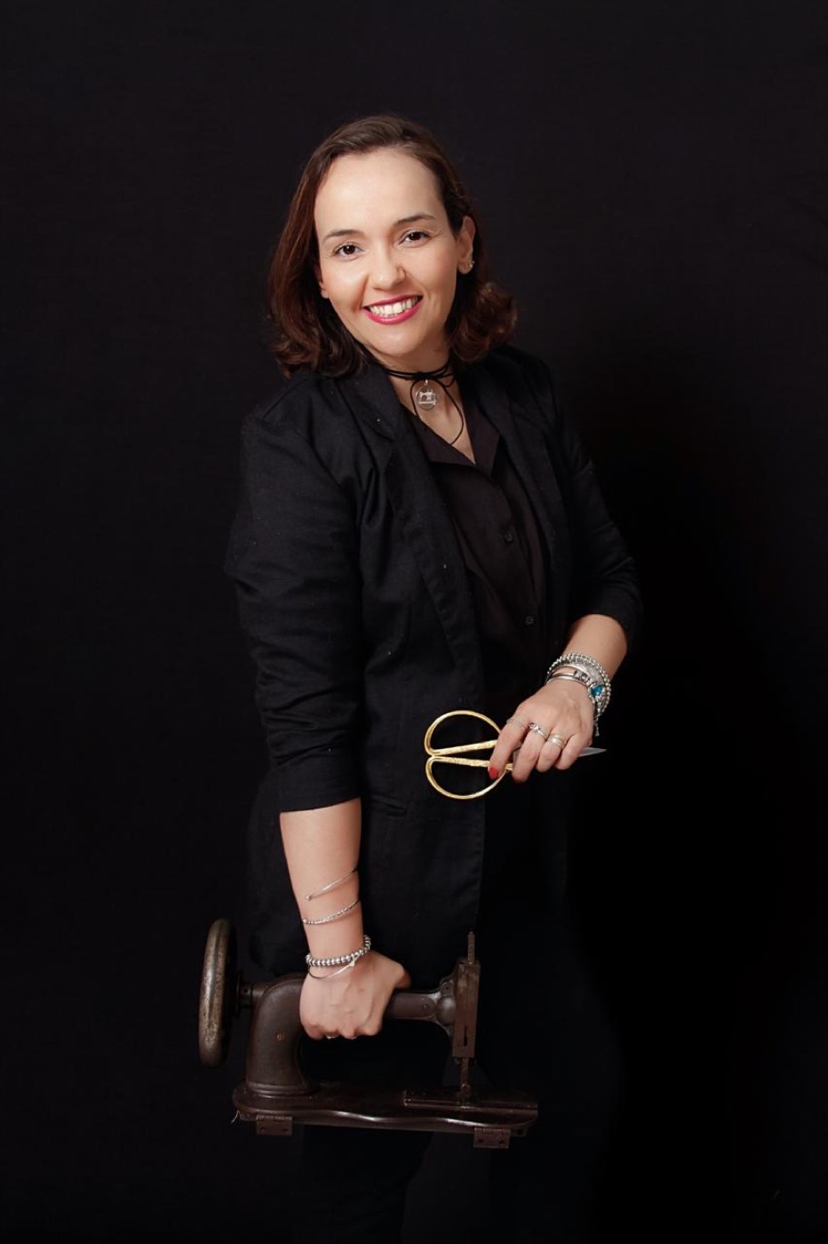 Fabiana Ribeiro de preto, segurando uma máquina de costura antiga e uma tesoura dourada